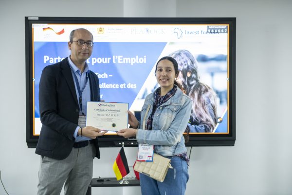 Un homme remet un « Certificate of Achievement » à une jeune femme devant un écran affichant le logo d’Invest for Jobs, à côté d’un drapeau allemand de table.