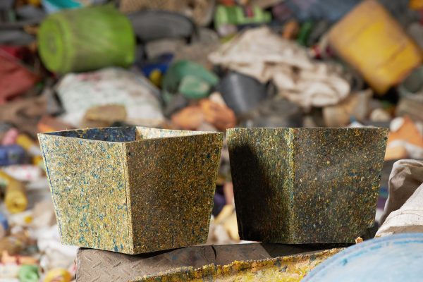 Two square planters made of pressed, multicoloured recycled plastic, with plastic waste blurred in the background.