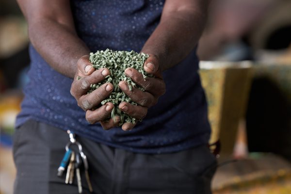 Two hands hold a handful of green recycled plastic pellets, with a planter blurred in the background.