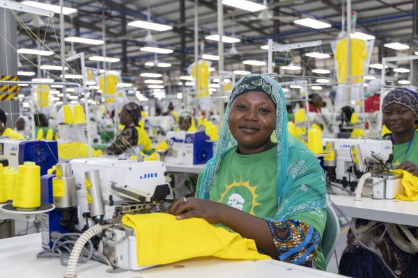 A smiling seamstress in a headscarf sews yellow fabric at her machine, with more workstations of a large garment factory behind her.