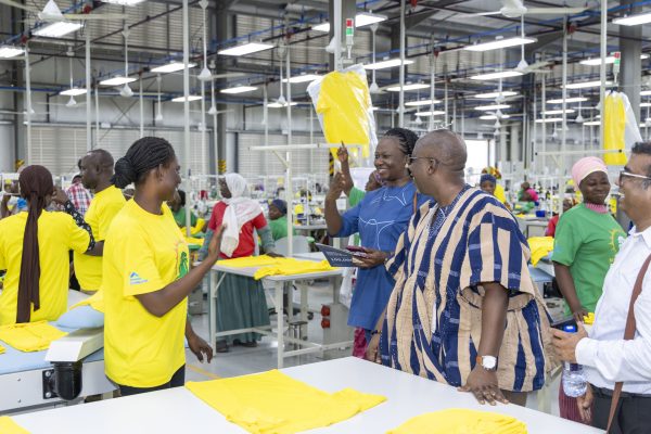 Visitors are shown around a garment factory; a smiling worker holds up a yellow T-shirt, with more yellow garments laid out on the tables.
