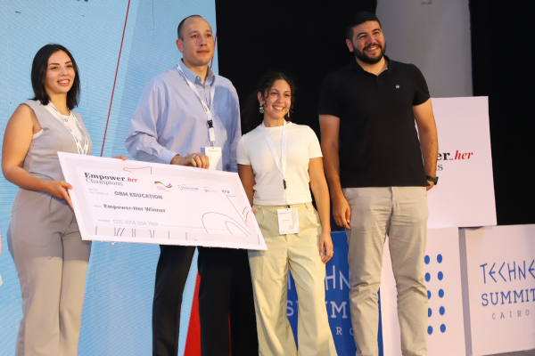 Four people on stage at the Techne Summit Cairo; one woman holds a large “Empower Her Winner” award cheque for OBM Education.