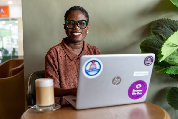 Une femme souriante aux lunettes vertes est assise dans un café devant un ordinateur portable portant l’autocollant « Because She Can », à côté d’un verre de latte macchiato.