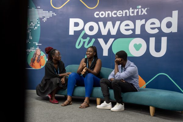 Trois jeunes personnes discutent sur un canapé devant un mur portant le slogan de Concentrix « Powered by You ».