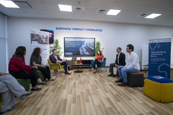 Panel discussion in a meeting room with the wall slogan “Empower through knowledge”: seven people sit in a semicircle in front of a screen, with German and Moroccan table flags and an “Opportunités de croissance au Maroc” roll-up banner.