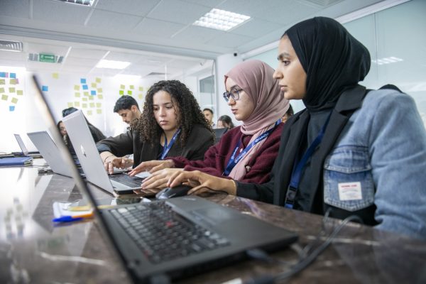 Des jeunes travaillent avec concentration sur des ordinateurs portables à une longue table, avec des post-it au mur en arrière-plan.