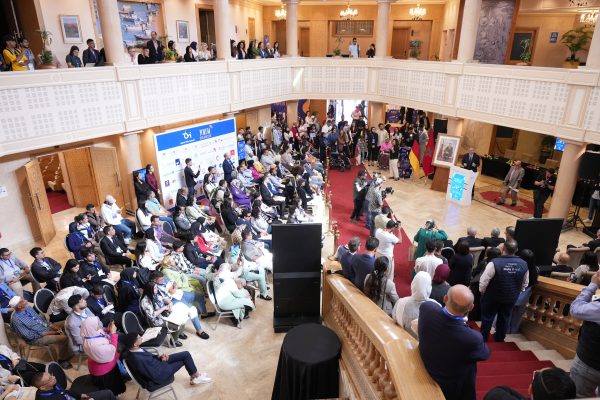 View from a gallery over a packed hall where a large audience follows a speech at a lectern, beside German and Moroccan flags.