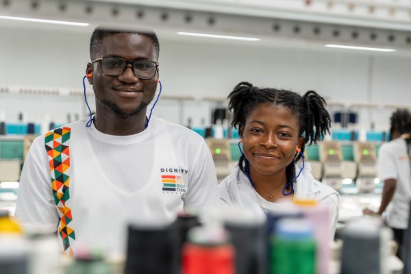 Un jeune homme et une jeune femme sourient à l’objectif, avec des machines à broder en arrière-plan et des bobines de fil floues au premier plan.