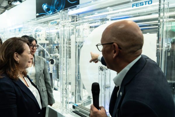 Ein Festo-Vertreter mit Mikrofon zeigt auf ein transparentes Automatisierungsexponat, mehrere Frauen hören zu.