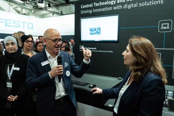 A guide with a microphone talks with a woman in front of a Festo display on control technology, with a group of visitors behind them.