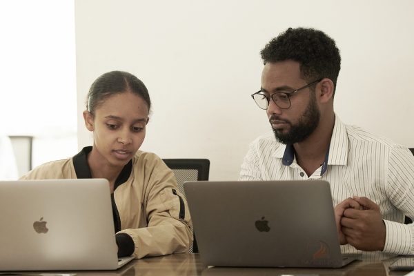 Eine junge Frau und ein Mann mit Brille sitzen nebeneinander an Laptops und schauen gemeinsam auf ihren Bildschirm.